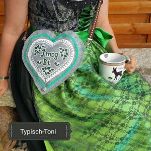 May include: A traditional German dirndl dress with a green and black floral pattern. The dress features a heart-shaped purse with the text "I mog Di" and a white mug with a deer silhouette. The outfit includes a white blouse and a green ribbon.
