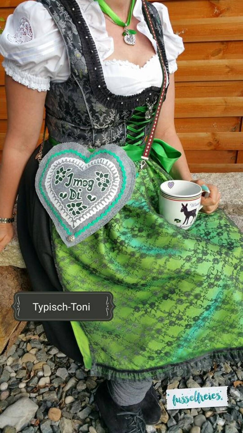 May include: A traditional German dirndl dress with a green and black floral pattern. The dress features a heart-shaped purse with the text "I mog Di" and a white mug with a deer silhouette. The outfit includes a white blouse and a green ribbon.