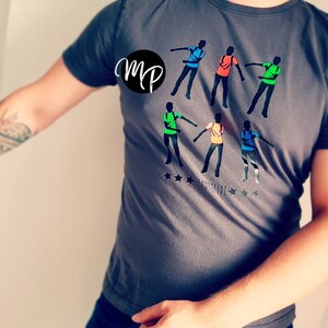 Könnte beinhalten: Ein graues T-Shirt mit einer Grafik von sechs tanzenden Personen. Die Grafik ist in verschiedenen Farben, darunter Grün, Blau, Orange und Rot. Der Text "MP" befindet sich in einem Kreis auf dem Shirt. Der Text "INSPIRIDE" befindet sich unter der Grafik.