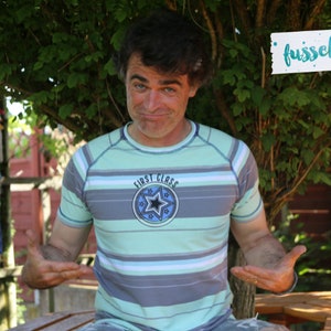 May include: A man wearing a light blue and gray striped shirt with a blue circle and a star on the front. The shirt says "First Class" on it. He is standing in front of a green bush and a wooden fence. The text "fussselfreies" is written on a white piece of paper.
