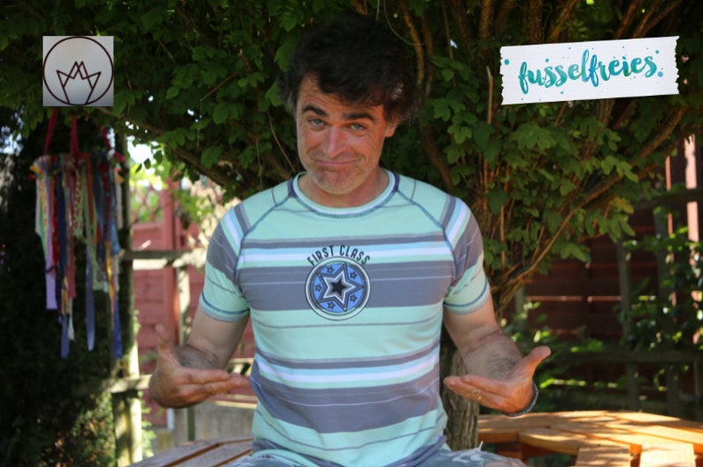 May include: A man wearing a light blue and gray striped shirt with a blue circle and a star on the front. The shirt says "First Class" on it. He is standing in front of a green bush and a wooden fence. The text "fussselfreies" is written on a white piece of paper.