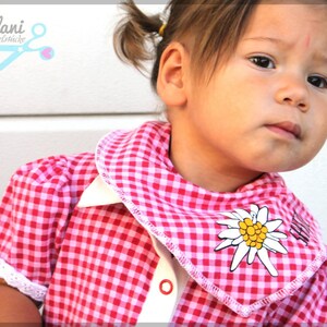 May include: A pink and white gingham shirt with a white collar and cuffs. The shirt has a decorative edelweiss flower on the collar. The image also includes the text "milla & lani" and a pair of scissors.