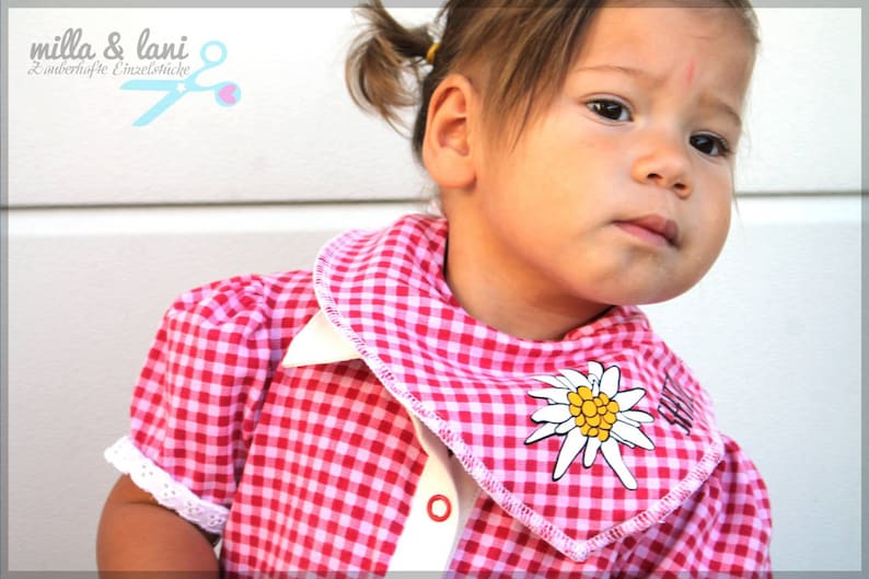 May include: A pink and white gingham shirt with a white collar and cuffs. The shirt has a decorative edelweiss flower on the collar. The image also includes the text "milla & lani" and a pair of scissors.