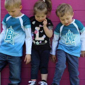 May include: Three children wearing blue and white shirts with a star and letter design. The shirts say "B", "L", and "M" on them. The children are standing in front of a pink wall with a white sign that says "fusselfreies."