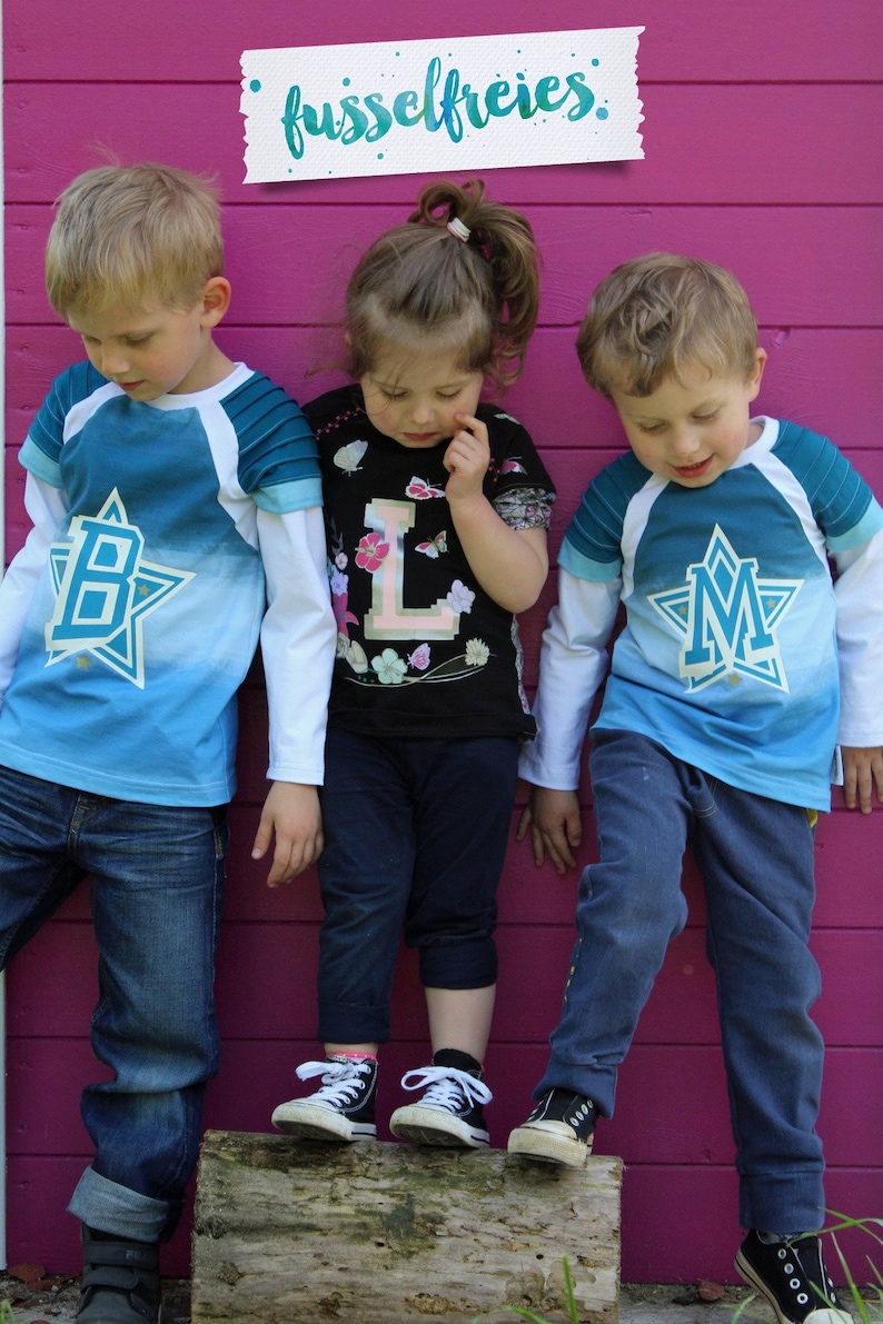 May include: Three children wearing blue and white shirts with a star and letter design. The shirts say "B", "L", and "M" on them. The children are standing in front of a pink wall with a white sign that says "fusselfreies."