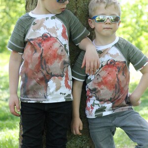 May include: Two children wearing green and white shirts with a brown bear graphic print. The shirts have a green and white striped sleeve. The children are wearing sunglasses and standing next to a tree. The text "Bobba Barchen" is visible on the tree.