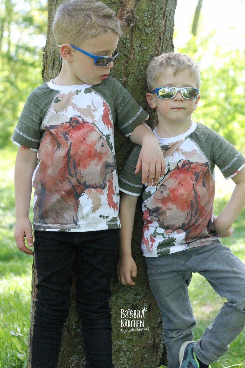May include: Two children wearing green and white shirts with a brown bear graphic print. The shirts have a green and white striped sleeve. The children are wearing sunglasses and standing next to a tree. The text "Bobba Barchen" is visible on the tree.