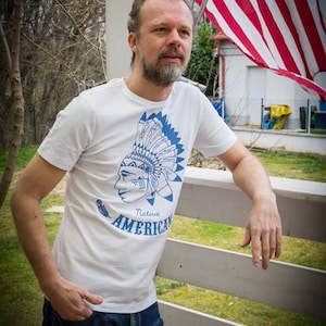 May include: A man wearing a white t-shirt with a blue graphic of a Native American headdress and the text "Native American". He is standing in front of a white wooden fence with green grass behind it. An American flag is waving in the background.