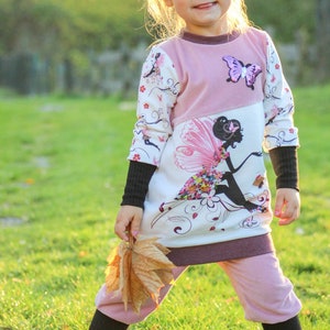 May include: A young girl wearing a pink and white long-sleeved dress with a fairy graphic and a butterfly applique. She is holding a brown leaf and smiling at the camera.