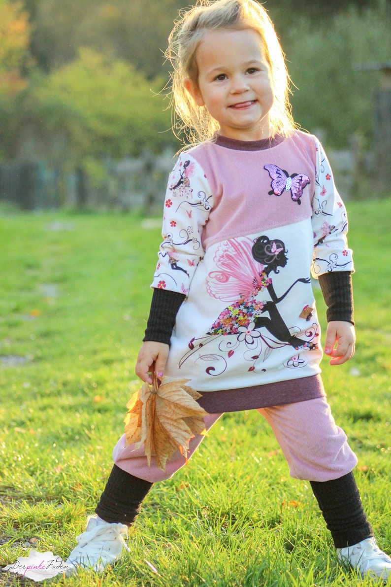May include: A young girl wearing a pink and white long-sleeved dress with a fairy graphic and a butterfly applique. She is holding a brown leaf and smiling at the camera.