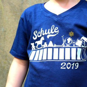 May include: A blue t-shirt with a white graphic design that says "Schule 2019". The graphic features a school desk with various school supplies, including a rocking horse, a globe, a backpack, and a lightbulb.