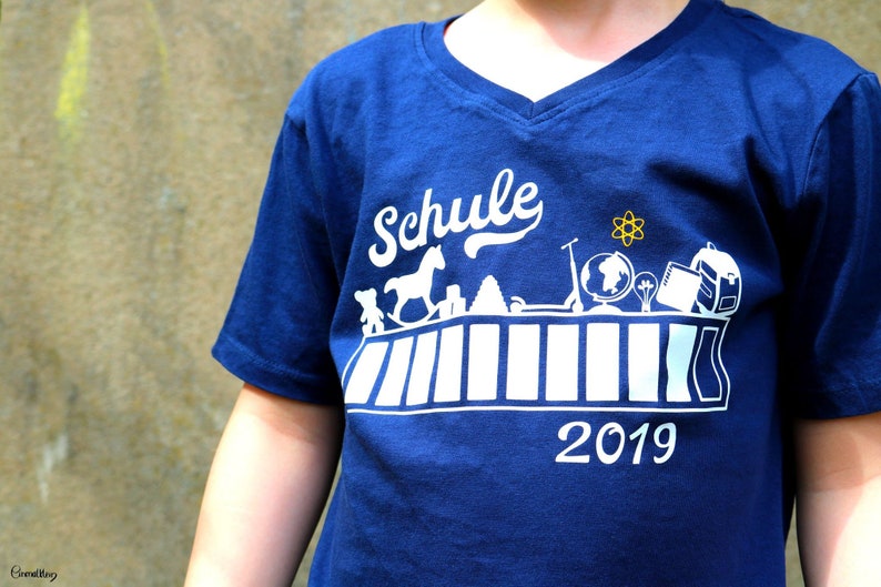 May include: A blue t-shirt with a white graphic design that says "Schule 2019". The graphic features a school desk with various school supplies, including a rocking horse, a globe, a backpack, and a lightbulb.