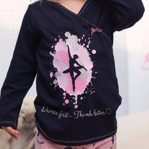 Op de afbeelding: Een zwart shirt met lange mouwen met een roze en zilveren glitterontwerp van een ballerina in een cirkel. De tekst "Dance first - Think later" is in zilveren glitter onder het ontwerp geschreven.