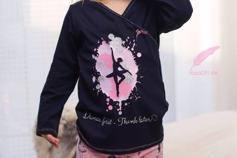 Op de afbeelding: Een zwart shirt met lange mouwen met een roze en zilveren glitterontwerp van een ballerina in een cirkel. De tekst "Dance first - Think later" is in zilveren glitter onder het ontwerp geschreven.