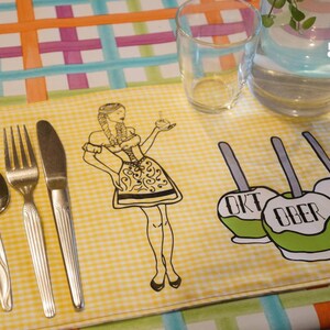 May include: A yellow gingham placemat with a black and white illustration of a woman in traditional German attire, and three green candy apples with the words "OKT OBER FEST". Silverware includes a spoon, fork, and knife.