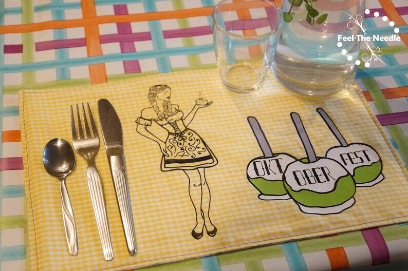 May include: A yellow gingham placemat with a black and white illustration of a woman in traditional German attire, and three green candy apples with the words "OKT OBER FEST". Silverware includes a spoon, fork, and knife.