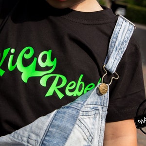 May include: A black t-shirt with a green graphic that says "Kica Rebel" on the front. The shirt is being worn by a child wearing denim overalls.
