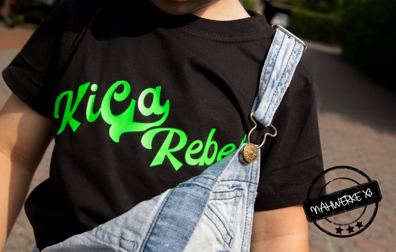 May include: A black t-shirt with a green graphic that says "Kica Rebel" on the front. The shirt is being worn by a child wearing denim overalls.