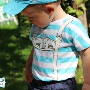 May include: A child's t-shirt with horizontal white and turquoise stripes. The shirt features a decorative design resembling lederhosen suspenders, with a central emblem of a deer and foliage. The child is also wearing a blue cap.