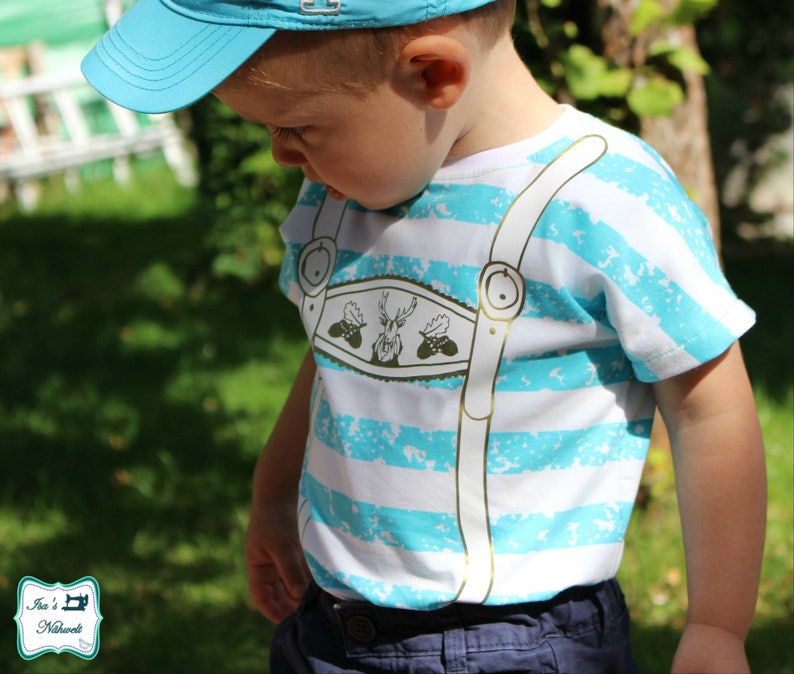 May include: A child's t-shirt with horizontal white and turquoise stripes. The shirt features a decorative design resembling lederhosen suspenders, with a central emblem of a deer and foliage. The child is also wearing a blue cap.