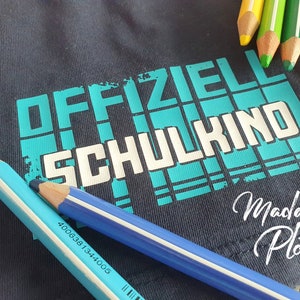 May include: A dark blue t-shirt with the text "OFFIZIELL SCHULKIND" in white letters on a turquoise background. The text "Madame Plott" is written in cursive below the main text.
