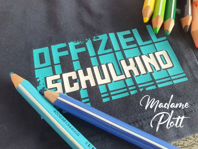 May include: A dark blue t-shirt with the text "OFFIZIELL SCHULKIND" in white letters on a turquoise background. The text "Madame Plott" is written in cursive below the main text.