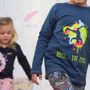 Op de afbeelding: Twee kinderen dragen shirts met lange mouwen met grafische ontwerpen. Het shirt van de jongen is donkerblauw met een gele, groene en rode afbeelding van een persoon die een dansbeweging maakt en de tekst "Feel the Beat". Het shirt van het meisje is zwart met een roze afbeelding van een dansende persoon.