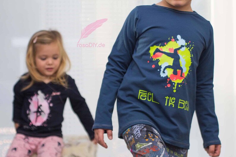 Op de afbeelding: Twee kinderen dragen shirts met lange mouwen met grafische ontwerpen. Het shirt van de jongen is donkerblauw met een gele, groene en rode afbeelding van een persoon die een dansbeweging maakt en de tekst "Feel the Beat". Het shirt van het meisje is zwart met een roze afbeelding van een dansende persoon.