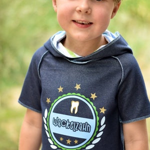 May include: A young boy wearing a blue denim hoodie with a green, white, and gold emblem that says "Wackelzahn".