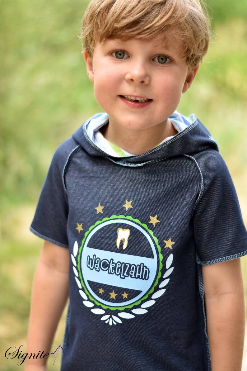 May include: A young boy wearing a blue denim hoodie with a green, white, and gold emblem that says "Wackelzahn".