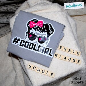 May include: Gray t-shirt with a black and white graphic of a girl with pink sunglasses and a pink bow. The text "#COOLGIRL" is printed in white below the graphic. The words "ERSTE KLASSE SCHULE" are spelled out in white letters on wooden tiles.