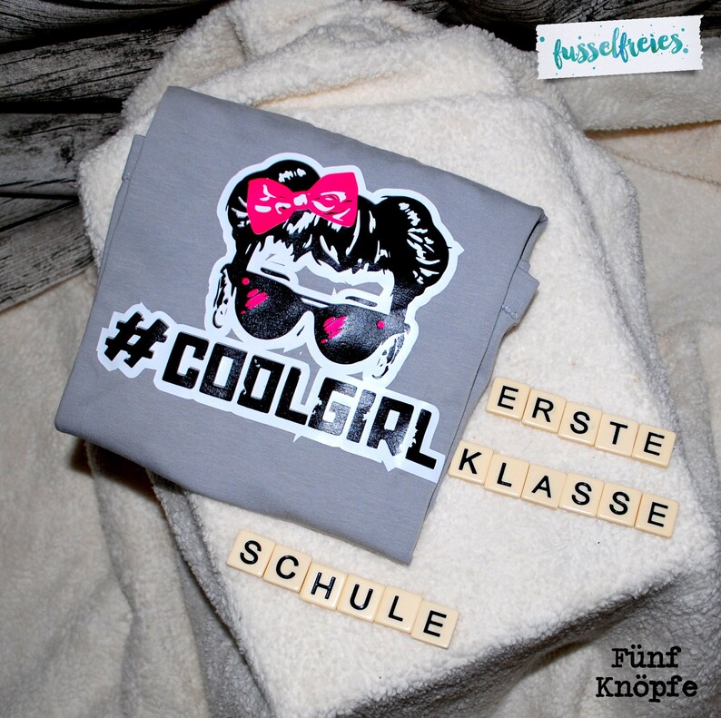 May include: Gray t-shirt with a black and white graphic of a girl with pink sunglasses and a pink bow. The text "#COOLGIRL" is printed in white below the graphic. The words "ERSTE KLASSE SCHULE" are spelled out in white letters on wooden tiles.