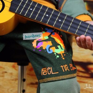 Op de afbeelding: Een kind draagt een groen shirt met een kleurrijke grafische print en de tekst "Feel the Beat" en speelt een gele ukulele. Het shirt heeft ook een klein wit label met de tekst "fusselfreies".