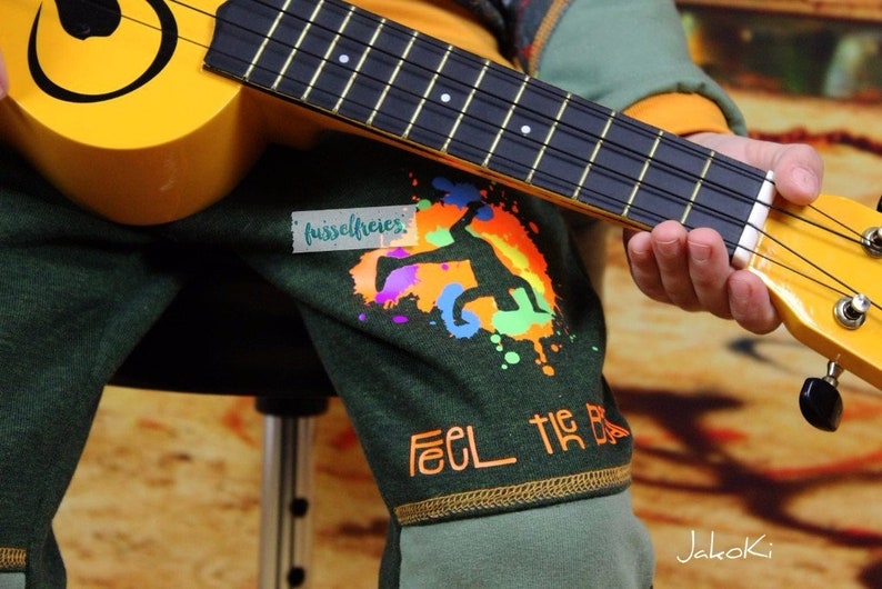 Op de afbeelding: Een kind draagt een groen shirt met een kleurrijke grafische print en de tekst "Feel the Beat" en speelt een gele ukulele. Het shirt heeft ook een klein wit label met de tekst "fusselfreies".