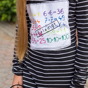 May include: A black and white striped long-sleeved shirt with a white patch on the front. The patch has colorful handwritten math equations, including "1x1=1", "6x6=36", "7x7=49", "3x3=9", "4x4=16", "5x5=25", "9x9=81", and "10x10=100". The patch also has a red rectangle with the word "Erledigt" written in it.