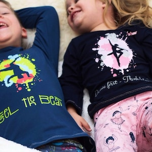 Könnte beinhalten: Zwei Kinder tragen langärmelige Shirts mit bunten Grafikdesigns. Das Shirt des Jungen sagt "Feel the Beat" und zeigt eine Silhouette eines Tänzers. Das Shirt des Mädchens hat ein pinkfarbenes und silbernes Sprühdesign mit einer Ballerina-Silhouette und dem Text "Dance first - Think later".