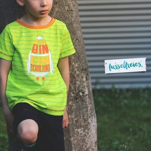 May include: A green and orange striped t-shirt with a rocket ship graphic and the text "Bin ein Schulkind". The shirt has a yellow trim around the neck and sleeves.