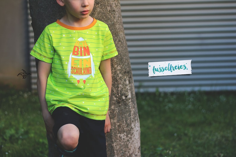 May include: A green and orange striped t-shirt with a rocket ship graphic and the text "Bin ein Schulkind". The shirt has a yellow trim around the neck and sleeves.