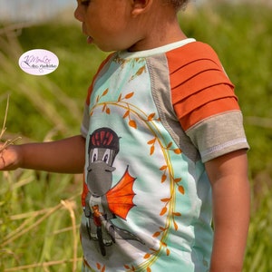 May include: A light blue t-shirt with a grey and orange design. The shirt features a cartoon dragon riding a motorcycle. The text "das Nadelstudio" is visible on the shirt.