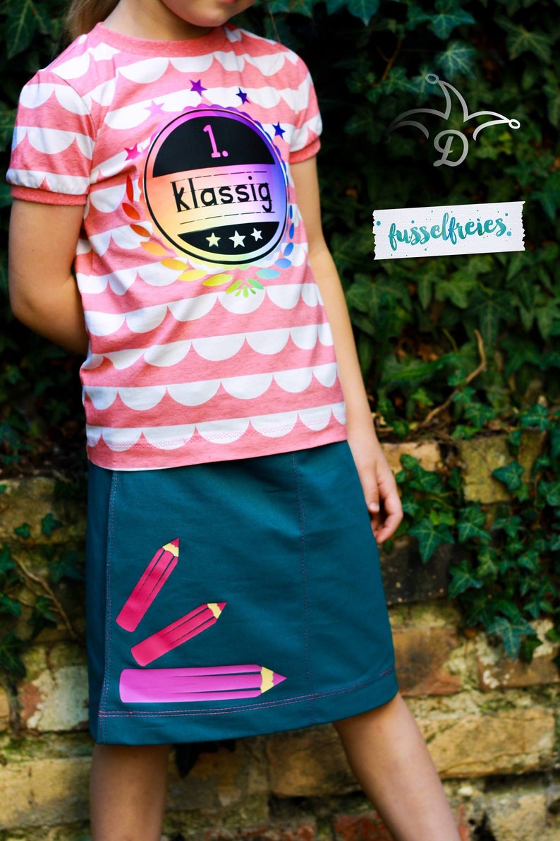 May include: A young girl wearing a pink and white striped shirt with a rainbow-colored patch that says "1.  klassig" and a teal skirt with pink pencil designs.
