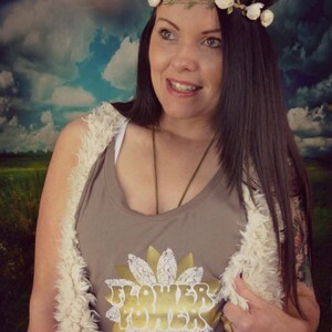 May include: A woman wearing a brown tank top with a gold sunflower graphic and the text "Flower Power". She is wearing a white faux fur vest and a flower crown.
