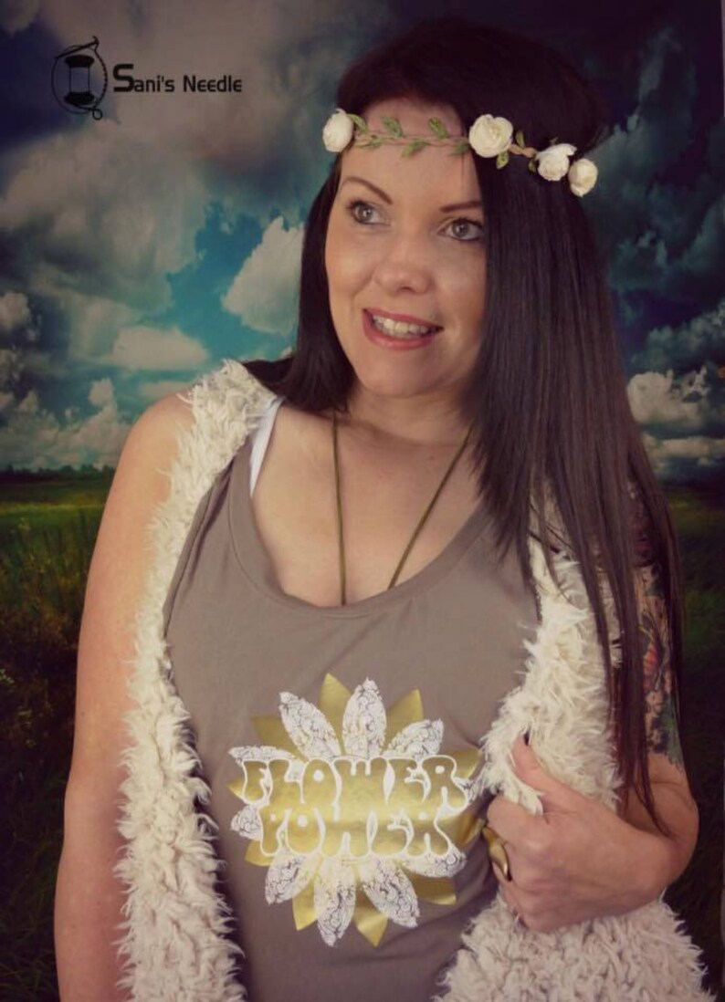 May include: A woman wearing a brown tank top with a gold sunflower graphic and the text "Flower Power". She is wearing a white faux fur vest and a flower crown.