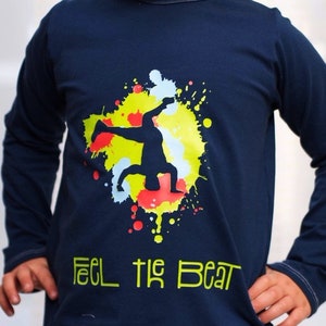 Op de afbeelding: Een marineblauw longsleeve shirt met een kleurrijk verfspatten ontwerp met een silhouet van een persoon die breakdancet. De tekst "Feel the Beat" is in groen onder het ontwerp gedrukt.