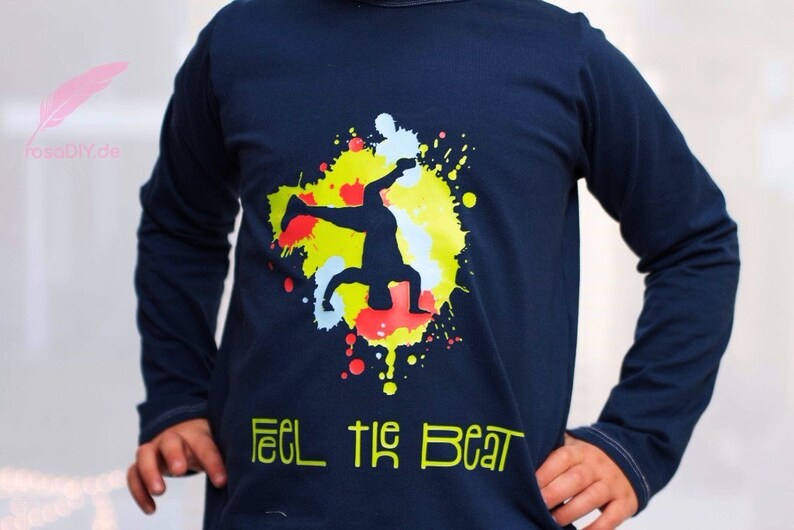 Op de afbeelding: Een marineblauw longsleeve shirt met een kleurrijk verfspatten ontwerp met een silhouet van een persoon die breakdancet. De tekst "Feel the Beat" is in groen onder het ontwerp gedrukt.