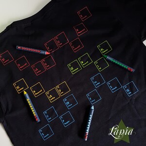 May include: A black t-shirt with a periodic table printed on it. The table is filled with colorful squares, each containing the symbol and atomic number of a chemical element. The t-shirt is surrounded by colorful markers.