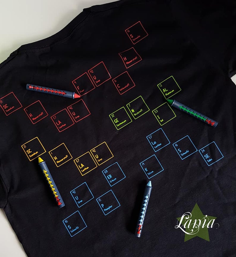 May include: A black t-shirt with a periodic table printed on it. The table is filled with colorful squares, each containing the symbol and atomic number of a chemical element. The t-shirt is surrounded by colorful markers.