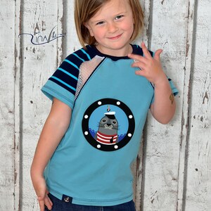 May include: A young person wearing a light blue t-shirt with a striped sleeve and a mesh panel. The t-shirt has a round applique of a seal wearing a sailor hat and a red and white striped shirt. The seal is looking out of a porthole with waves around it. The person is also wearing a dark blue denim skirt with an anchor on the front.