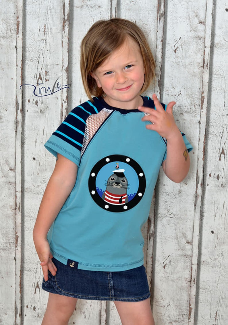 May include: A young person wearing a light blue t-shirt with a striped sleeve and a mesh panel. The t-shirt has a round applique of a seal wearing a sailor hat and a red and white striped shirt. The seal is looking out of a porthole with waves around it. The person is also wearing a dark blue denim skirt with an anchor on the front.