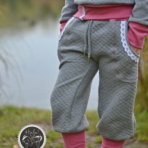 May include: Gray sweatpants with a quilted pattern and pink trim. The sweatpants have a white lace trim and a drawstring waist. The model is wearing pink leg warmers and gray shoes.