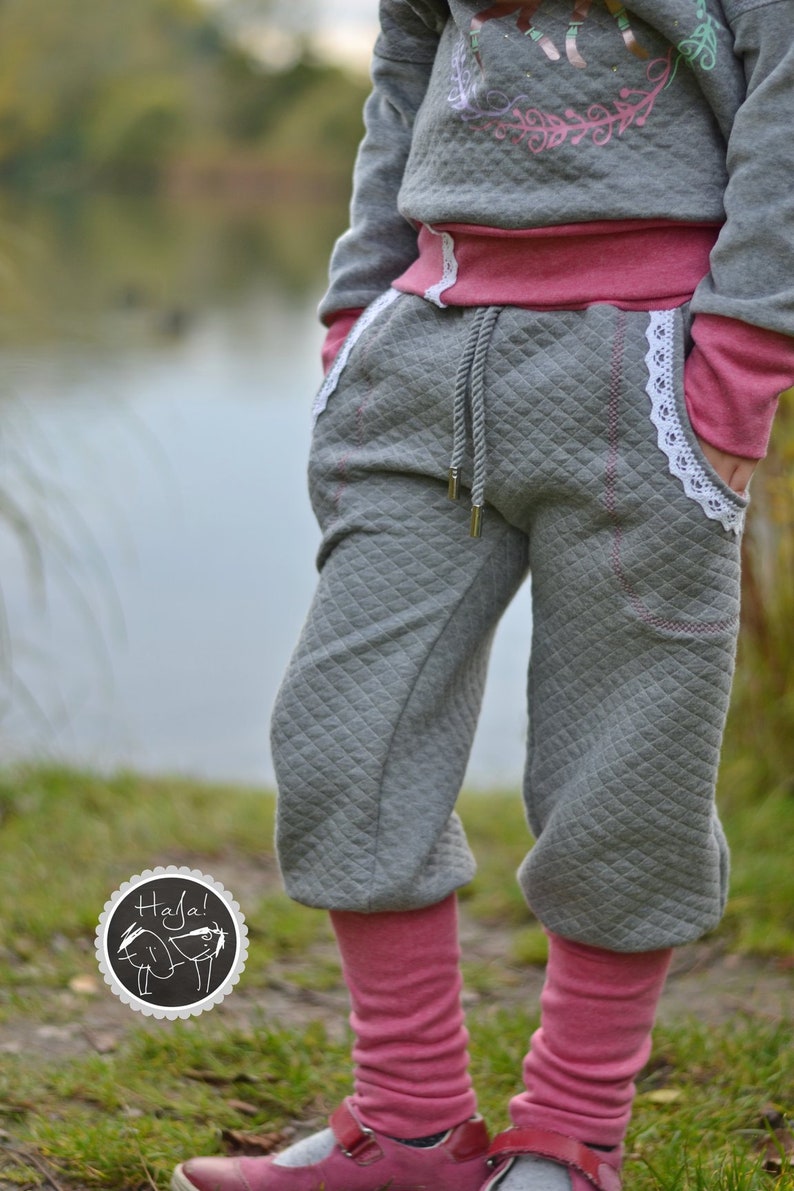 May include: Gray sweatpants with a quilted pattern and pink trim. The sweatpants have a white lace trim and a drawstring waist. The model is wearing pink leg warmers and gray shoes.
